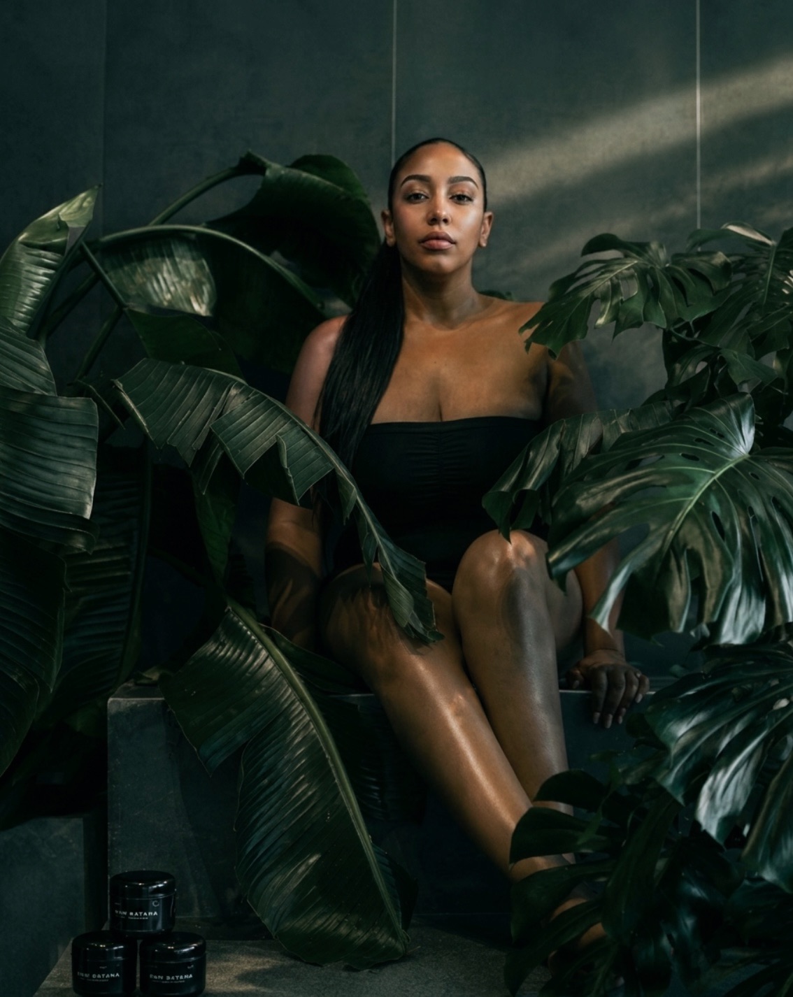Editorial portrait seated among rainforest leaves with Raw Batana jars